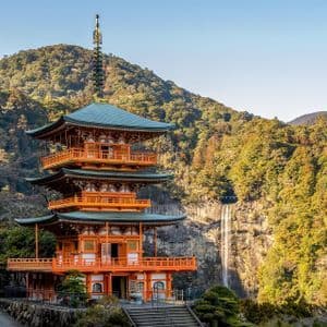 Una pagoda giapponese arancione a tre piani si erge su un fianco di montagna, con una grande cascata e una fitta foresta alle sue spalle.