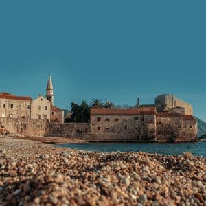 Una vista dal basso da una spiaggia di ciottoli che guarda verso una storica città di pietra con un campanile di chiesa sulla costa.