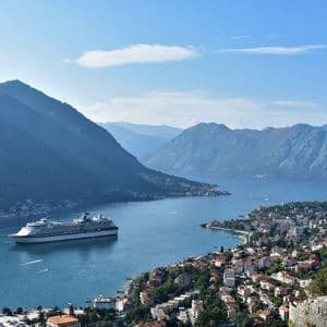 Una grande nave da crociera naviga in una baia vicino a una città costiera, circondata da ripide montagne sotto un cielo blu.