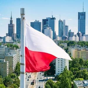 La bandera de Polonia ondea en primer plano con un horizonte urbano moderno que presenta rascacielos y exuberantes árboles verdes en el fondo.