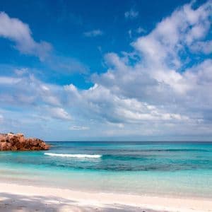 Ein tropischer Strand mit weißem Sand und türkisfarbenem Wasser schmiegt sich an eine felsige Küste mit Palmen unter einem blauen, bewölkten Himmel.