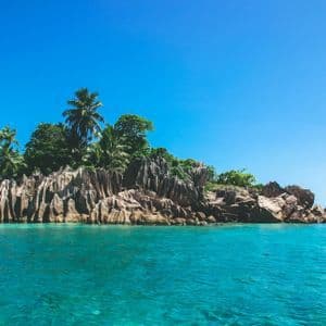 Eine kleine, felsige Insel, bewachsen mit üppig grünen Palmen, umgeben von ruhigem türkisfarbenem Wasser unter einem klaren blauen Himmel.