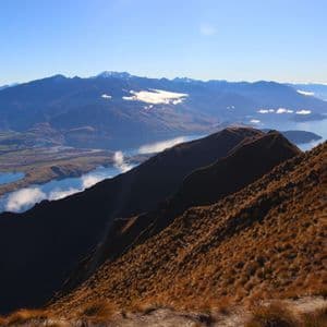Une crête montagneuse herbeuse mène à un sommet où deux randonneurs contemplent un vaste paysage de lacs et de montagnes sous un ciel bleu.
