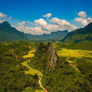Vue aérienne d'une route sinueuse traversant une vallée luxuriante remplie de forêts et de rizières, entourée de montagnes sous un ciel bleu.