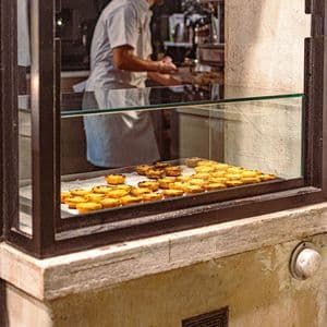 Una vetrina di una pasticceria piena di pastéis de nata, con un fornaio al lavoro sullo sfondo.