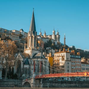 Un pont rouge enjambe une rivière devant une église gothique et une ville à flanc de colline, sous un ciel bleu clair.