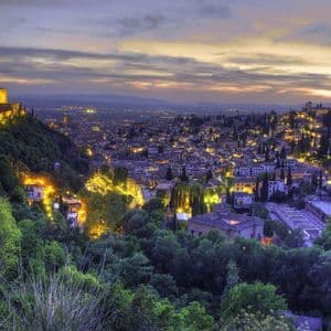Una fortaleza iluminada en una colina boscosa domina una extensa ciudad con luces parpadeantes al anochecer.