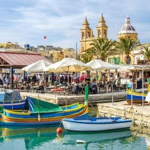 Barche colorate sono ormeggiate in un porto accanto a vivaci caffè sul lungomare, con una città storica e una chiesa sullo sfondo.