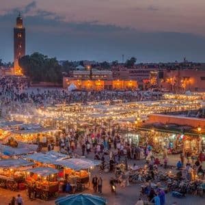 Una vista dall'alto di una piazza cittadina affollata al tramonto, piena di bancarelle illuminate e un alto minareto sullo sfondo.