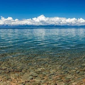 Una vista panoramica di un lago limpido con fondale roccioso, montagne innevate in lontananza e un cielo azzurro brillante con nuvole.