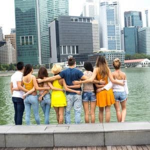 A WeRoad group trip seen from behind, standing with arms around each other looking out at a modern city skyline across the water.