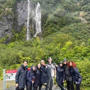 Un groupe WeRoad, vêtus d'imperméables, pose pour une photo devant une haute cascade dans une vallée verdoyante.