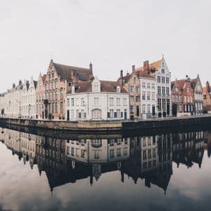 Una hilera de edificios históricos con tejados a dos aguas se alza a lo largo de un canal, con sus fachadas perfectamente reflejadas en el agua tranquila bajo un cielo nublado.
