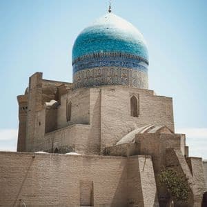 Un edificio in mattoni color sabbia con una grande cupola piastrellata di blu si erge in un cortile pavimentato sotto un cielo sereno.