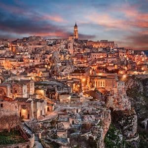 Un'antica città di pietra incastonata in una scogliera, illuminata da luci calde sotto un suggestivo cielo al tramonto.