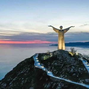 Una statua illuminata con le braccia distese si erge su una cima di montagna che si affaccia sul mare al tramonto, con un sentiero tortuoso e illuminato sottostante.