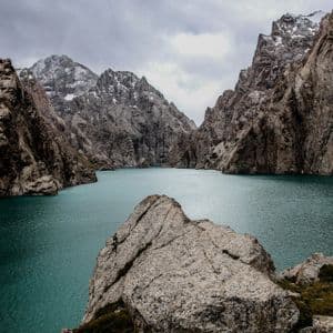 Un lago turchese incastonato tra montagne ripide e rocciose con cime innevate sotto un cielo nuvoloso.