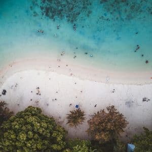 Vista aérea de personas nadando en agua turquesa cristalina y relajándose en una playa de arena blanca con árboles verdes.