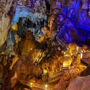 Un escalier en bois serpente à travers une grande grotte avec des formations rocheuses colorées illuminées par des lumières bleues et jaunes, avec des bateaux sur l'eau en contrebas.