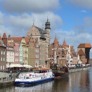Edificios coloridos e históricos con tejados a dos aguas bordean un muelle fluvial donde varios barcos están atracados bajo un cielo parcialmente nublado.