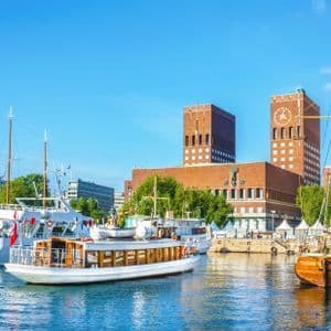 Diverse barche e yacht sono ormeggiati in un porto cittadino, di fronte a un grande edificio in mattoni rossi con due torri, sotto un cielo azzurro.