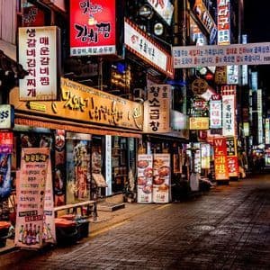 A paved city street at night, lined with buildings covered in colorful, glowing neon signs written in Korean.