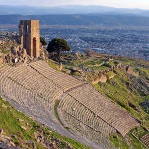 Un antiguo anfiteatro de piedra construido en una ladera cubierta de hierba, con ruinas y una ciudad moderna de fondo.