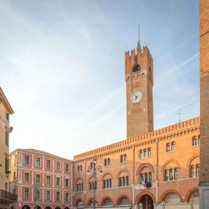 Una alta torre de reloj de ladrillo rojo se alza sobre una plaza histórica con edificios abovedados bajo un cielo brillante.