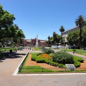 Un grande parco cittadino in una giornata di sole, con viali pavimentati, un monumento bianco e un lontano palazzo rosa circondato da alberi.