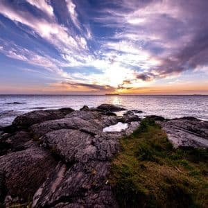 Una costa rocciosa ed erbosa si affaccia sull'oceano al tramonto, con un cielo pieno di nuvole striate viola e blu.