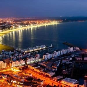 Vue aérienne nocturne d'une ville côtière lumineuse, avec des lumières éclairant une longue plage incurvée et un port.