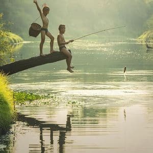 Deux garçons sur une bûche enjambant une rivière : l'un pêche à la canne et attrape un poisson bondissant, tandis que l'autre célèbre derrière lui avec un panier.