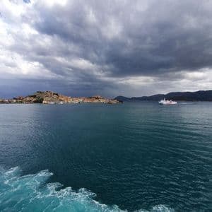 Vista dal mare di una località costiera su una collina e un traghetto che naviga sotto un cielo cupo e nuvoloso.