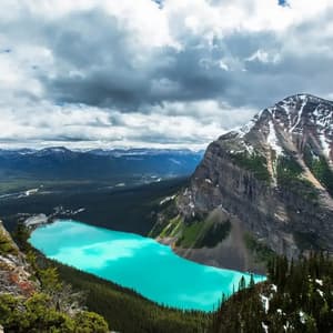 A high-angle view of a bright turquoise lake surrounded by pine forests and a large, snow-patched mountain under a cloudy sky.