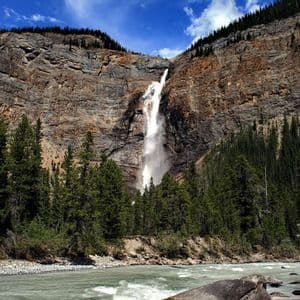 Una alta cascada cae por un acantilado de roca estratificada hacia un bosque de pinos, con un río ancho y caudaloso en primer plano.