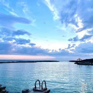 Eine Metallleiter führt in eine ruhige, klare blaue Bucht, umgeben von Felsen unter einem bewölkten Himmel in der Dämmerung.
