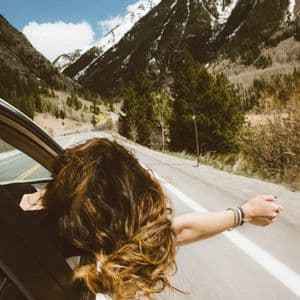 Una persona con cabello largo y castaño se asoma por la ventanilla de un coche en movimiento con el brazo extendido, mientras recorre una carretera de montaña.