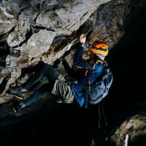 Una persona con un casco naranja y una linterna frontal escala la pared rocosa dentro de una cueva oscura.