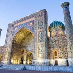 Une médersa ornée de mosaïques complexes bleues et dorées, une grande arche, un dôme turquoise et des minarets, illuminée au crépuscule.