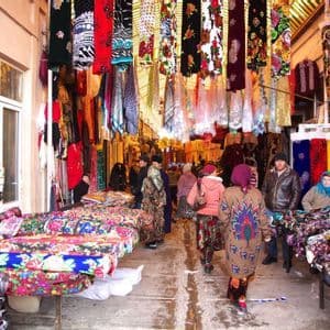 A busy outdoor market aisle crowded with people browsing stalls filled with colorful textiles and fabrics.