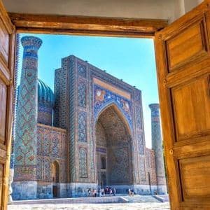 An ornate building with intricate blue and gold mosaic tiles and a dome, viewed from inside through a set of open wooden doors.