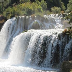 Ein breiter, mehrstufiger Wasserfall stürzt über felsige Absätze in einen sonnenbeschienenen Fluss, umgeben von üppigem Grün.