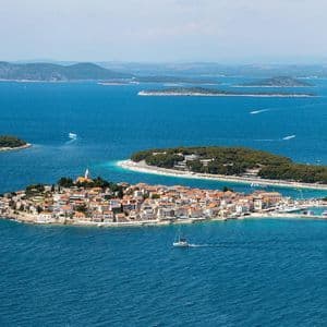 Luftaufnahme einer Küstenstadt auf einer kleinen Halbinsel, umgeben von blauem Meer, mit segelnden Booten und weiteren Inseln im Hintergrund.