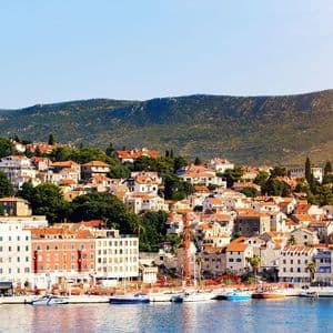 Eine Küstenstadt mit orangefarbenen Ziegeldächern und weißen Gebäuden schmiegt sich an einen grünen Hügel, mit Booten, die im blauen Hafen darunter vor Anker liegen.