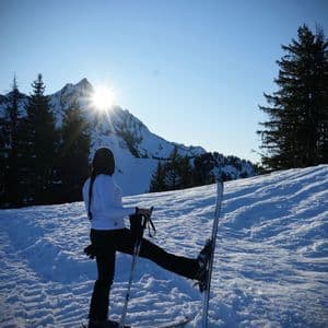 A person on skis stretches on a snowy mountain with the sun shining over a distant peak.