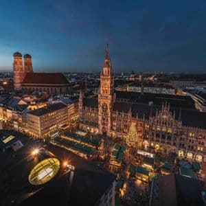 Una veduta aerea di un vivace mercatino di Natale in una piazza storica al tramonto, con edifici gotici illuminati sotto un cielo blu scuro.