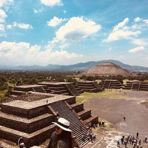 Une personne coiffée d'un chapeau blanc contemple un ancien site archéologique avec de grandes pyramides en pierre sous un ciel partiellement nuageux.