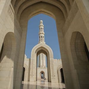 A view through multiple light-colored stone arches of an ornate mosque, with a tall minaret visible against a clear blue sky.