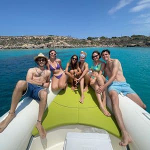 Un gruppo WeRoad di sei persone in costume da bagno sorride a prua di una barca, galleggiando su acqua turchese vicino a una costa rocciosa.