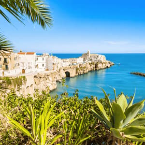 Una città costiera con edifici bianchi, arroccata su una scogliera rocciosa che domina un mare blu calmo, incorniciata da piante verdi e foglie di palma.